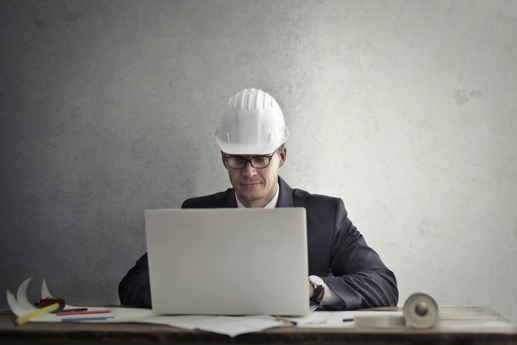 Professional engineer wearing hardhat working on construction plans at desk with laptop in office.